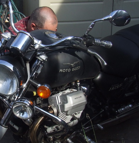 A man squats beyond a matt black & chrome Guzzi Jackal, an Italian V twin shaft-driven motorbike